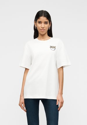 Hosszú sötét hajú nő fehér, díszített logóval ellátott pólóban és sötétkék farmerral, egy egyszerű háttér előtt állva.