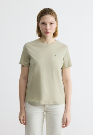 Γυναίκα με κοντά ξανθά μαλλιά φοράει μπεζ κοντομάνικη μπλούζα και λευκό παντελόνι, στέκεται μπροστά σε ένα απλό φωτεινό φόντο και κοιτάζει προς τα εμπρός.