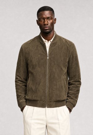 Uomo che indossa una giacca bomber in suede verde oliva sopra una polo bianca con pantaloni plissettati color avorio, mani nelle tasche, sfondo neutro.