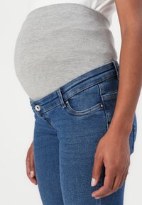 Maternity gray jersey belly band worn over blue denim jeans. Close-up on the side with a hand resting on the hip. Simple and functional design.