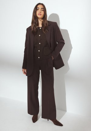 Blazer marrón ajustado y pantalones de pierna ancha, combinados con un top de punto marrón oscuro con botones y bolsillos, todo sobre un fondo blanco.