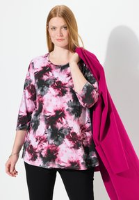 Halenka s květinovým vzorem v růžové a černé barvě, s tříčtvrtinovými rukávy a kulatým výstřihem. Modelka drží magentový kabát. Černé kalhoty.