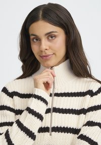 Krémový sveter s čiernymi horizontálnymi prúžkami, vysokým golierom a zipsom. Pletená textúra a rebrované manžety.