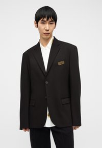 Νέος άνδρας με κοντά μαύρα μαλλιά φοράει ένα μαύρο, ραμμένο κοστούμι πάνω από ένα λευκό πουκάμισο με χρυσές λεπτομέρειες, σε απλό ανοιχτό φόντο.