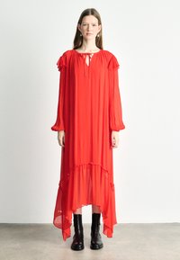 Largo vestido rojo con un corte holgado, mangas acampanadas, escote en forma de llave y dobladillo desigual. Combinado con botas negras hasta el tobillo. Tela suave y fluida.