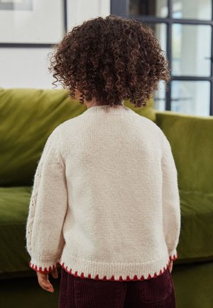 Otrok s kodrastimi lasmi, oblečen v krem pulover z rdečim robom, stoji v notranjosti z obrnjenim hrbtom proti kameri blizu zelene zofe.