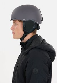 Grå hjälm med ventilationshål, justerbart hakband och vadderade öronskydd, kombinerad med en svart jacka med en logotyp på ärmen.