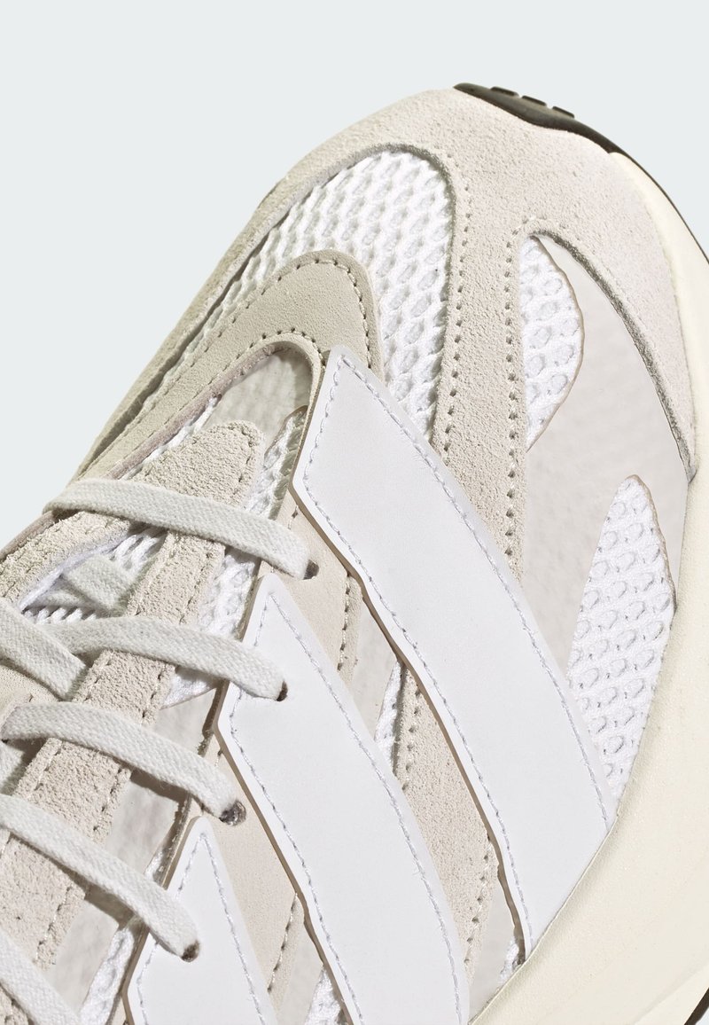 Sneaker aus einer Mischung von strukturierten Materialien, einschließlich Wildleder und Mesh in hellbeige und weiß, mit auffälligen weißen Streifen und Schnürsenkeln.