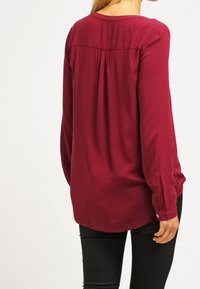 Blusa de manga larga color burdeos con escote redondo, pliegues sutiles en la parte trasera y detalles de botones en los puños. Tela suave y ligera.
