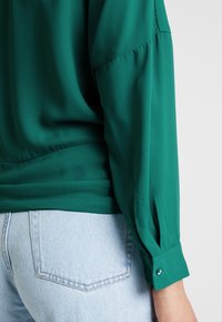 Türi langärmlige Bluse mit Knopfmanschetten, getragen mit hellblauen Jeans, die die Gesäßtasche und die Seitennaht zeigen.