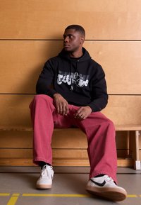 Felpa nera con logo, pantaloni rossi a gamba larga, sneakers bianche con dettagli neri. Il modello è seduto su una panchina di legno contro un muro di legno.