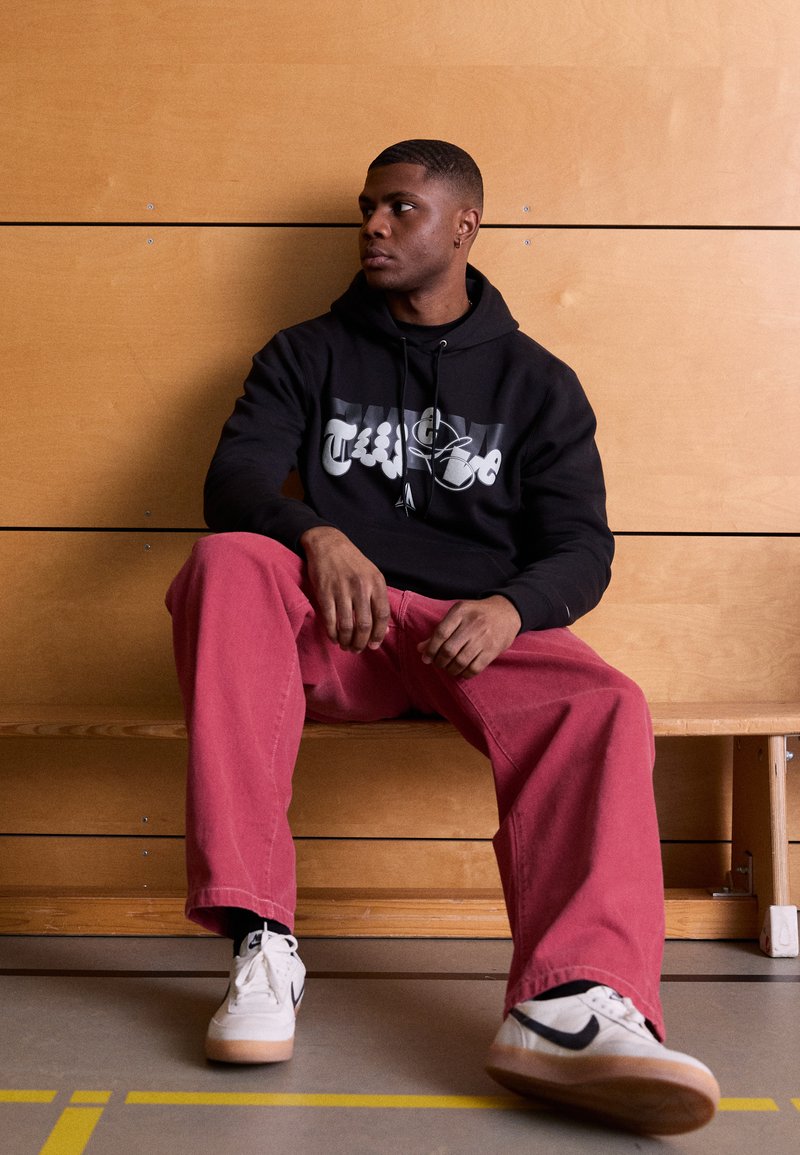 Czarna bluza z kapturem z logo, czerwone spodnie o szerokich nogawkach, białe sneakersy z czarnymi akcentami. Model siedzi na drewnianej ławce w pobliżu drewnianej ściany.