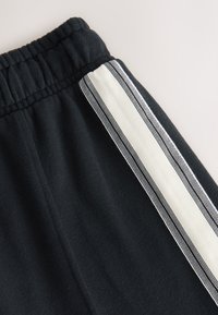 Schwarze Jogginghose mit weicher Textur, elastischem Bund und einem vertikalen weißen Streifen mit schwarzen Akzenten an der Seite.