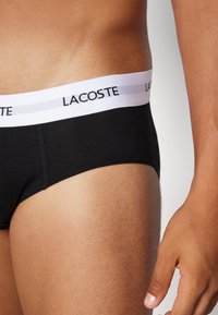 Schwarze Baumwollslips mit einem weißen elastischen Bund, der mit dem "LACOSTE"-Logo versehen ist. Nahaufnahme des Seiten- und Oberschenkelbereichs. Glatte Textur.
