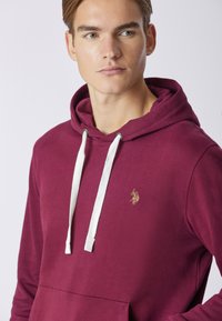 Felpa color bordeaux con cappuccio, dotata di un laccio bianco, presenta una tasca a marsupio e un logo ricamato in oro sul lato sinistro del petto, realizzata in tessuto morbido.