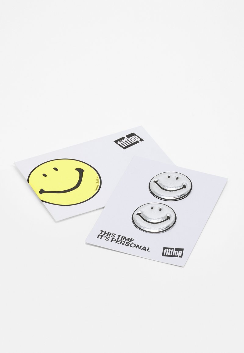 Duas cartas com designs de rostos sorridentes: um autocolante amarelo com contorno preto e outra com dois alfinetes circulares prateados num cartão branco.