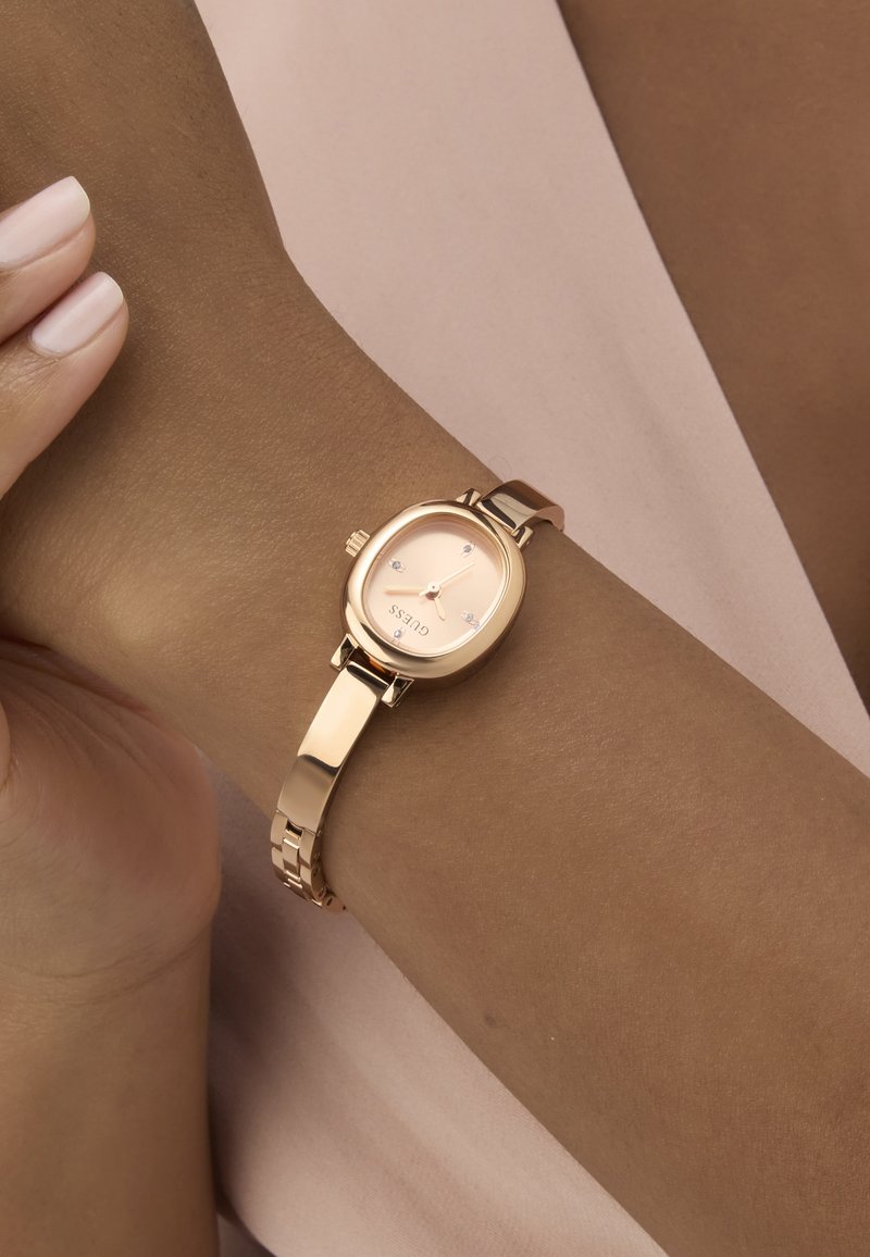 Aranyszínű, ovális tokú karóra, rózsaszín óralappal, három gyémántszerű órajellel és fémkapcsos szíjjal, amely egy bőrszínű csuklón van bemutatva.