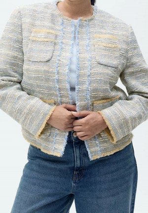 Person trägt eine strukturierte, hellfarbene, gewebte Jacke mit Fransen-Details über einem weißen Hemd und blauer Jeans, die Hände in der Taille verschränkt.