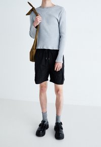 T-shirt gris clair à rayures, shorts noirs avec cordon de serrage, chaussettes grises et chaussures noires épaisses. Tenant un sac à bandoulière marron moyen.