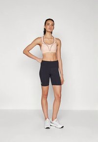 Hellrosa Sport-BH mit schwarzen Akzenten, kombiniert mit hochtaillierten schwarzen Radlerhosen. Model trägt weiße Sportschuhe. Glatte Stoffstruktur.