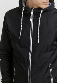Giacca leggera nera con cappuccio, cordini bianchi, due tasche con zip e cerniera a contrasto sul davanti. Texture del tessuto liscia.