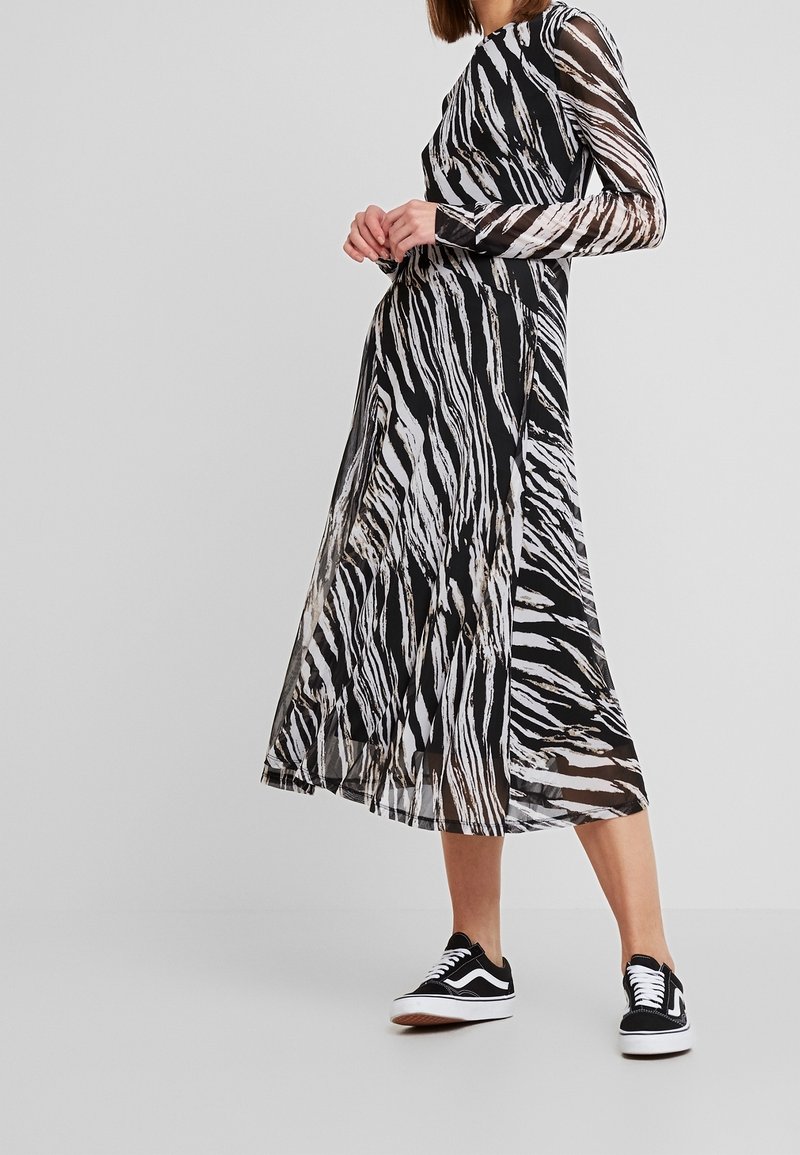 Femme portant une longue robe transparente noire et blanche à rayures zèbre avec des manches longues et des baskets noires avec des lacets blancs.