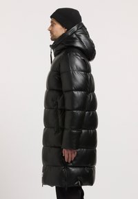 Cappotto puffer nero con cappuccio, design trapuntato, tasche laterali con zip e finitura lucida; indossato con un berretto lavorato a maglia nero.