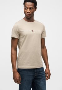 Μπεζ βαμβακερό T-shirt με στρογγυλή λαιμόκοψη, με λευκό κείμενο «TOMMY HILFIGER EST. 1985» και μικρό λογότυπο. Συνδυασμένο με σκούρο μπλε τζιν.