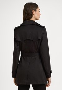 Czarny płaszcz przeciwdeszczowy z dwurzędowym zapięciem, szerokim kołnierzem i paskiem w talii. Posiada tylne karczki i klapy, wykonany z gładkiej tkaniny.