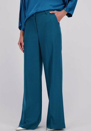 Pantalons large en teal en tissu lisse, présentant un devant plat et des poches latérales. Associés à des chaussures argentées à bout pointu.