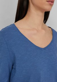 Donna che indossa una maglietta blu con scollatura a V, che mostra il collo, le clavicole e parte delle labbra su uno sfondo semplice.