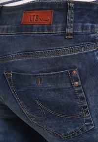 Tmavě modré džíny z denimu s hnědou koženou nášivkou označenou "LTB", prošívané detaily a dvě viditelné zadní kapsy s kovovými nýty.