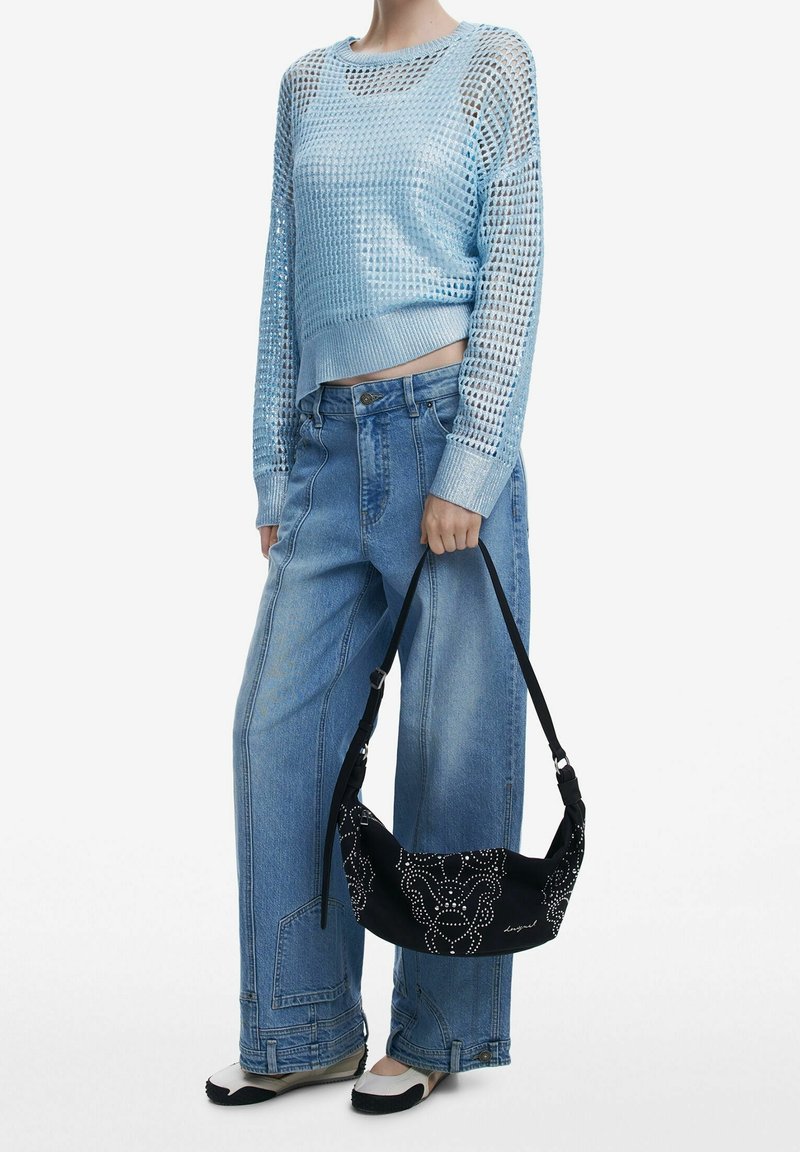 Personne portant un pull court en maille bleu clair et un jean à jambes larges, tenant un sac à main noir avec des motifs blancs en pointillés.