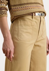 Tan cotton pants with a straight cut, featuring a back pocket and a silver belt buckle. The outfit is paired with a textured, patterned sweater.