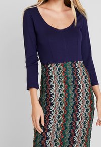 Granatowa bluzka z długim rękawem sparowana z dopasowaną spódnicą w wzory, z czerwonymi, zielonymi i kremowymi kwiatowymi motywami na ciemnym tle.