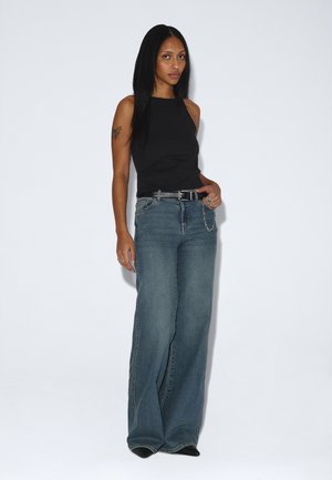 Donna con lunghi capelli neri che indossa una canottiera nera e jeans blu a gamba larga, in piedi davanti a uno sfondo bianco uniforme.