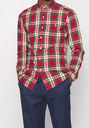 Mężczyzna ubrany w czerwoną, białą i zieloną koszulę w kratę z długim rękawem, zapiętą na guziki, oraz granatowe spodnie, ręce w kieszeniach, twarz niewidoczna.