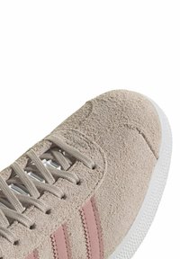 Bēša zamšādas sporta apavi ar rozā svītrām, teksturētu virspusi un pelēkām auklām. Balta gumijas zole ar protektora rakstu.