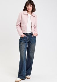 Donna che indossa una giacca rosa chiaro, una blusa bianca, jeans a gamba larga blu scuro e sneakers bianche, in piedi con le mani nelle tasche su uno sfondo bianco.