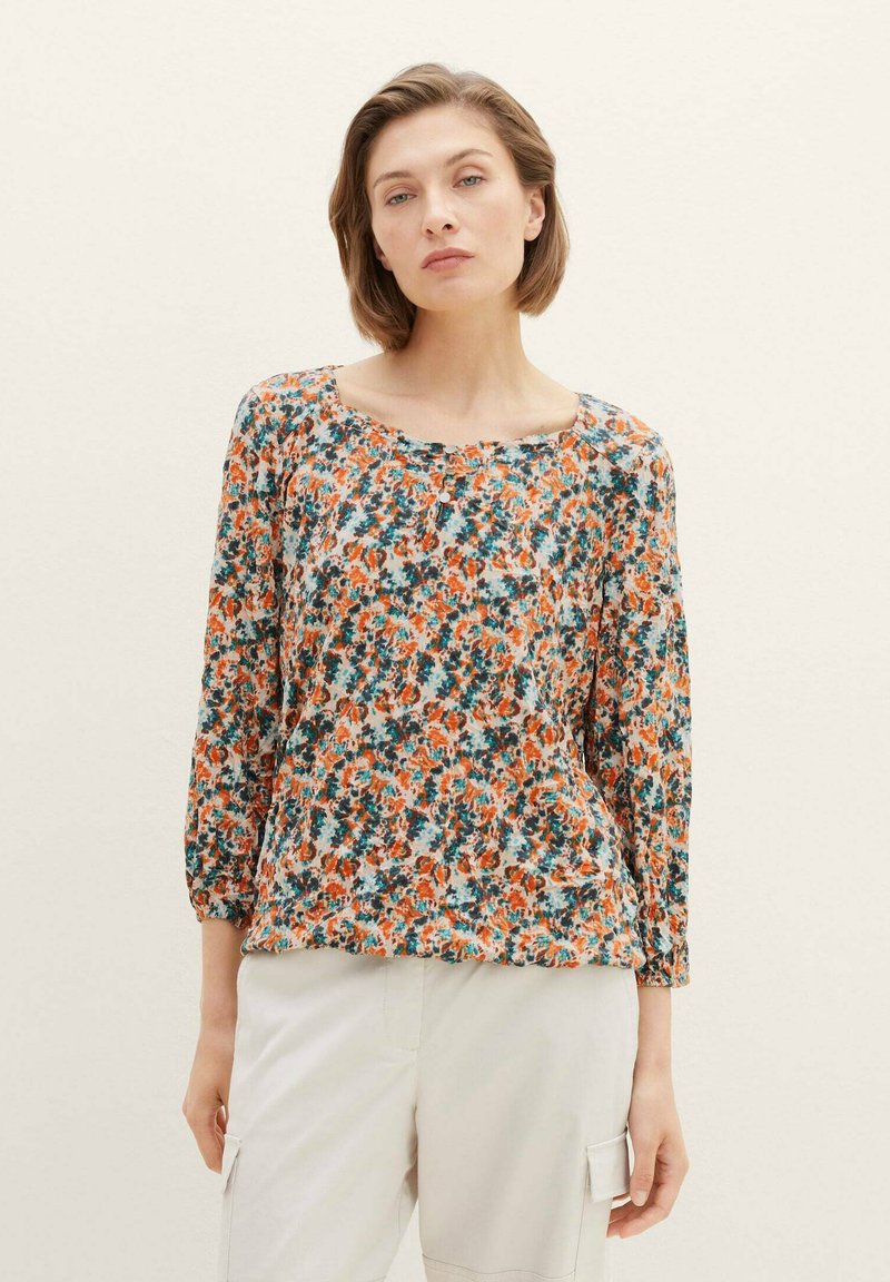 Blumenbluse mit langen Ärmeln, die eine Mischung aus orangefarbenen, türkisfarbenen und weißen Mustern aufweist, aus leichtem Stoff mit einem runden Ausschnitt gefertigt.