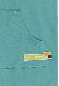 Il tessuto turchese presenta una tasca frontale con una cucitura a contrasto. Un'etichetta con il nome del marchio "loud + proud" è visibile, aggiungendo tocchi di colore.