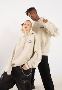 Beige hettegensere med frontlomme og snøring. Har svart logo-print. Sorte slitte jeans med kjededetalj. Minimalistisk design, uformell passform.