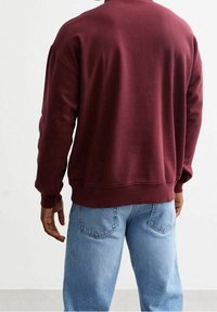 Felpa bordeaux con una vestibilità comoda e polsini a coste, abbinata a jeans in denim azzurro. La felpa presenta una texture liscia.