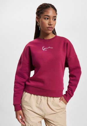 Burgundská mikina s kulatým výstřihem a vyšitým logem. Má volný střih, žebrované manžety a měkkou texturu. Nosená s béžovými kalhotami.