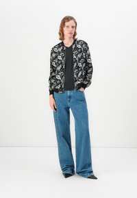 Veste bomber noire avec motif floral blanc, associée à un haut sombre et un jean bleu à jambes larges. Coupe slim, tissu lisse et fermeture éclair.