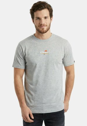 Homme aux cheveux bruns courts et à la barbe, portant un t-shirt gris clair à manches courtes avec un petit logo Ellesse au centre, debout devant un fond blanc.