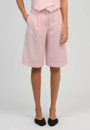 Femme portant un short ajusté rose clair arrivant aux genoux avec des poches latérales et des chaussures plates noires à bout pointu, debout devant un arrière-plan neutre.