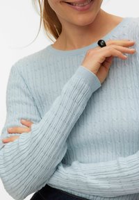 Pull bleu ciel en tricot avec une texture côtelée et un col rond. Les manches longues se terminent par des poignets ajustés. Une bague noire est portée à la main.