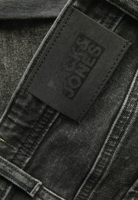 Crni traper tkanina s teksturiranom površinom; sadrži istaknutu crnu oznaku s natpisom "JACK & JONES" u uzdignutom pismu.