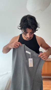 Maglione grigio lavorato a maglia senza maniche con scollo a V, caratterizzato da una trama a coste. Un'etichetta con il prezzo è attaccata al capo.